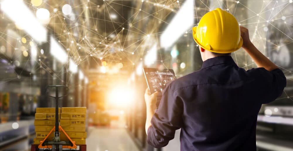 A man wearing a yellow hard hat reviews numbers on a tablet in an automated warehouse.